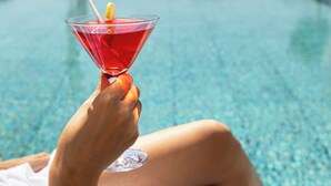 A person holding a red cocktail with a lemon slice, sitting by a pool with lounge chairs and a statue in the background. - Cassia Hotel Jerusalem (Jerusalem)