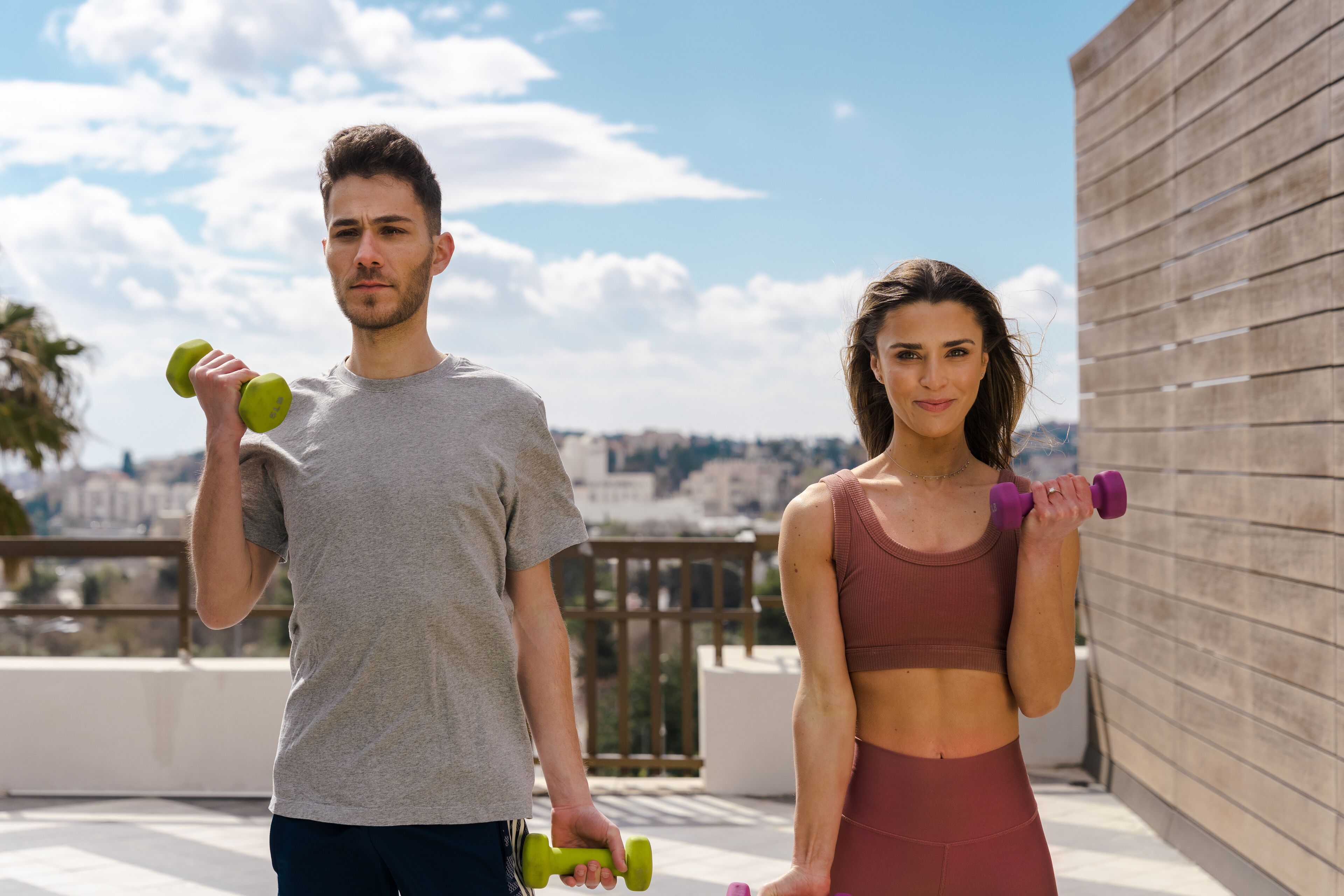 Two people exercising with dumbbells on a rooftop with a city view.