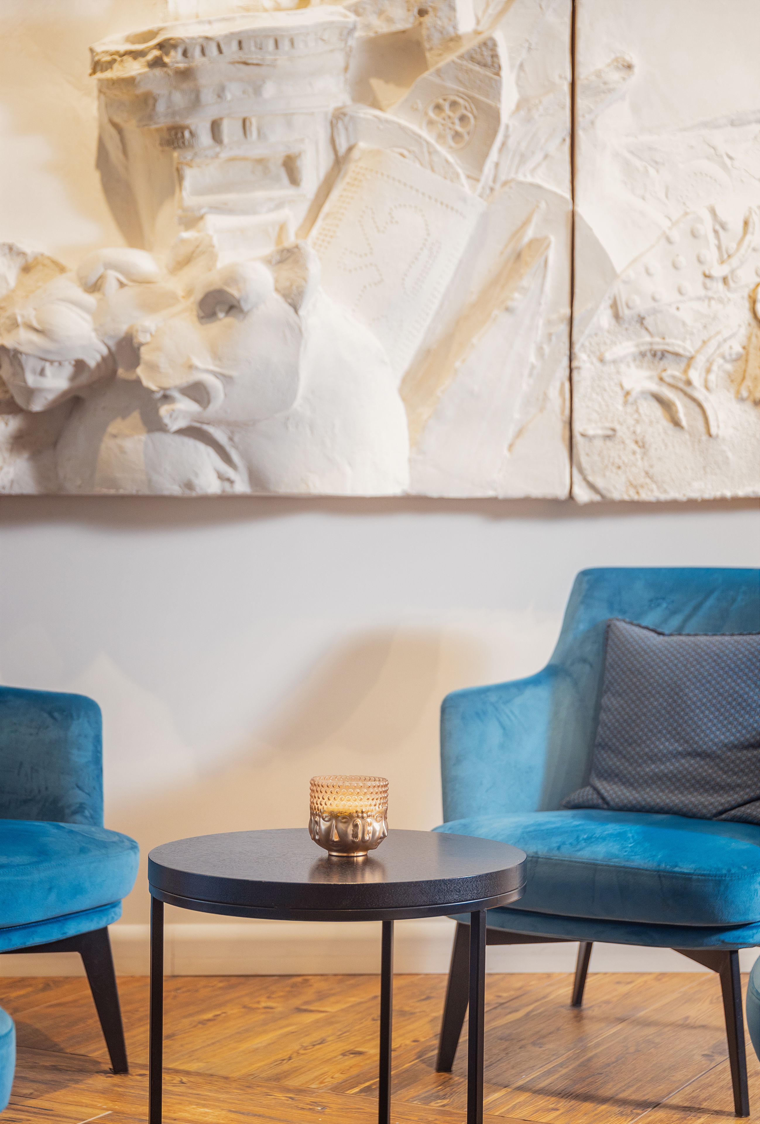 Een kamer met een ronde zwarte tafel, twee blauwe stoelen en een decoratief wandobject.