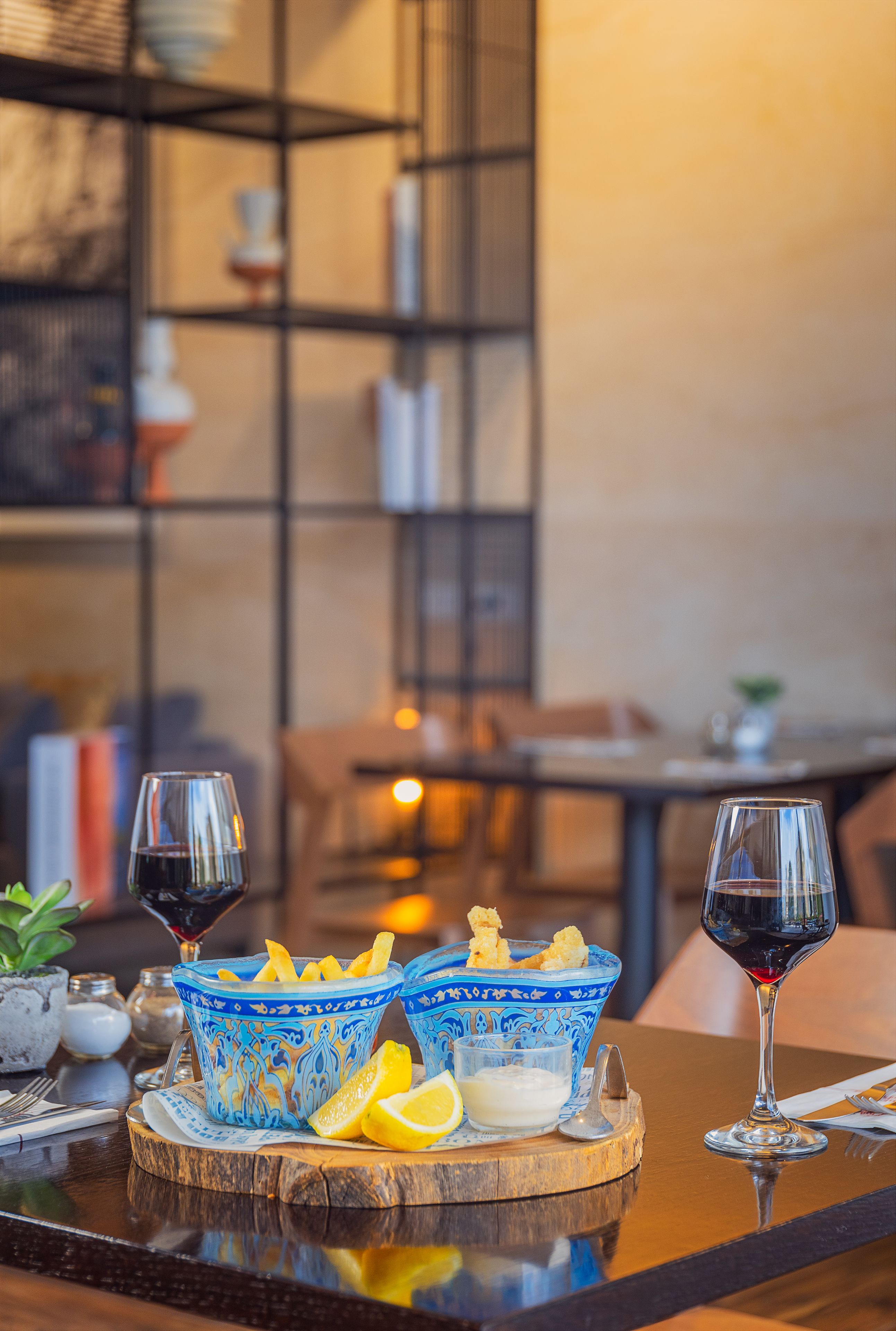 Una mesa de comedor con dos tazones azules estampados que contienen papas fritas, un gajo de limón y un recipiente pequeño con salsa. También hay dos copas de vino sobre la mesa.
