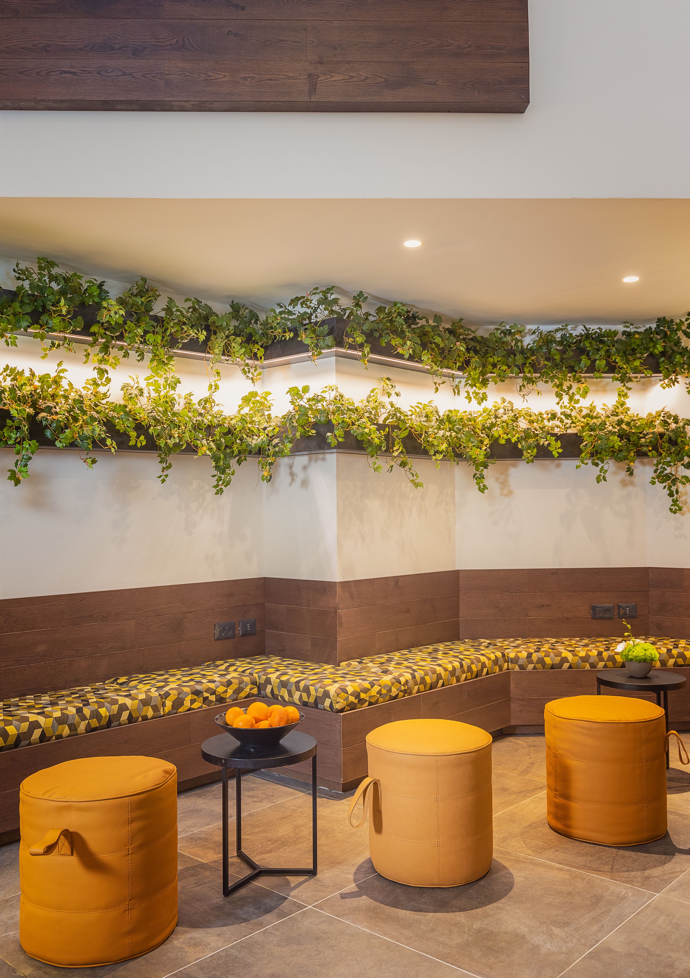A modern indoor seating area with cushioned benches and ottomans, a small table with a bowl of fruit, and a wall adorned with green climbing plants.