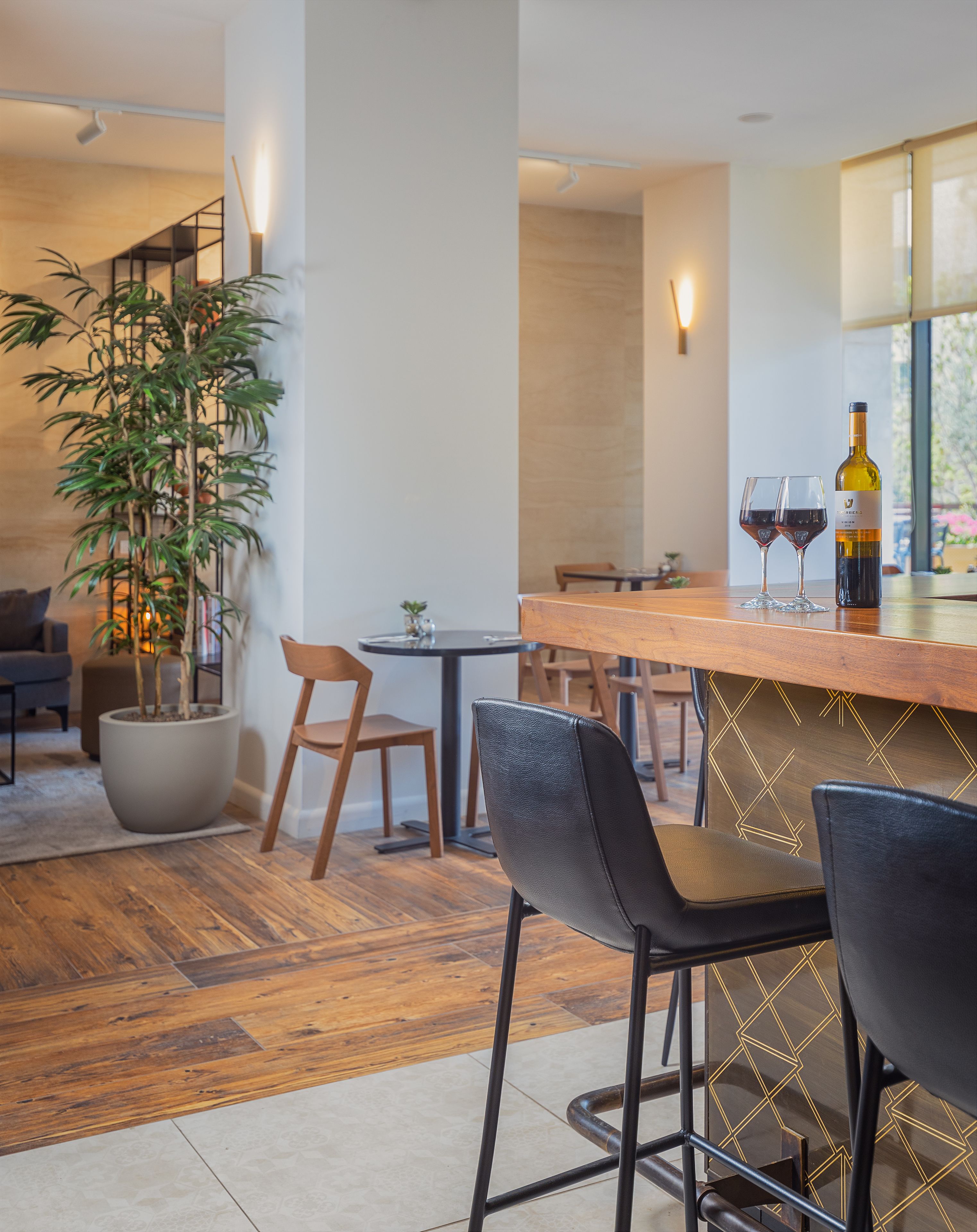 A modern bar area with a wooden counter, two chairs, a potted plant, and a bottle of wine on the bar.