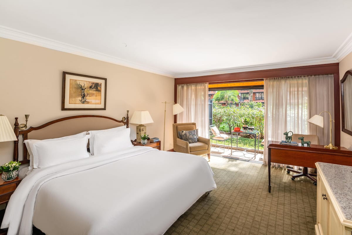 Quarto de hotel com cama, escrivaninha, cadeira e vista para um jardim.
