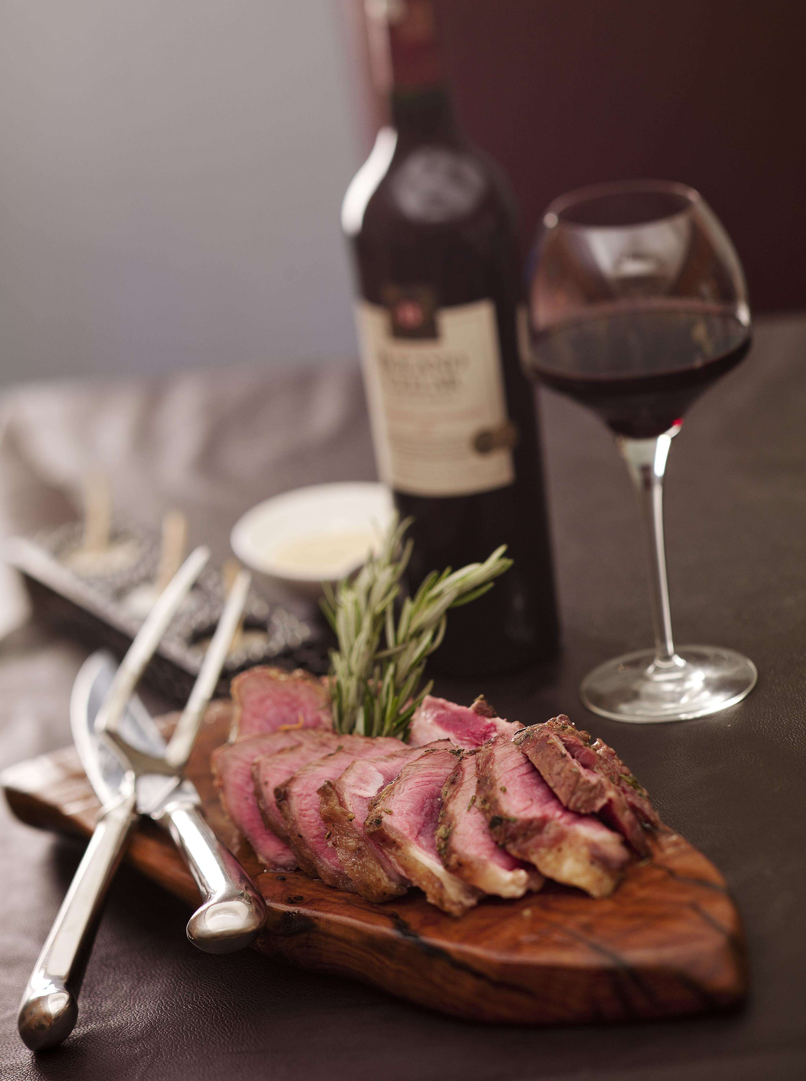 Una tabla de madera con carne cortada, una botella de vino y una copa de vino tinto sobre una mesa.