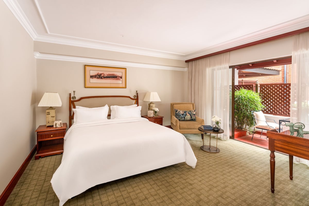 Quarto de hotel com uma cama grande, duas mesas de cabeceira com luminárias, uma cadeira, uma mesinha e vista para o exterior.