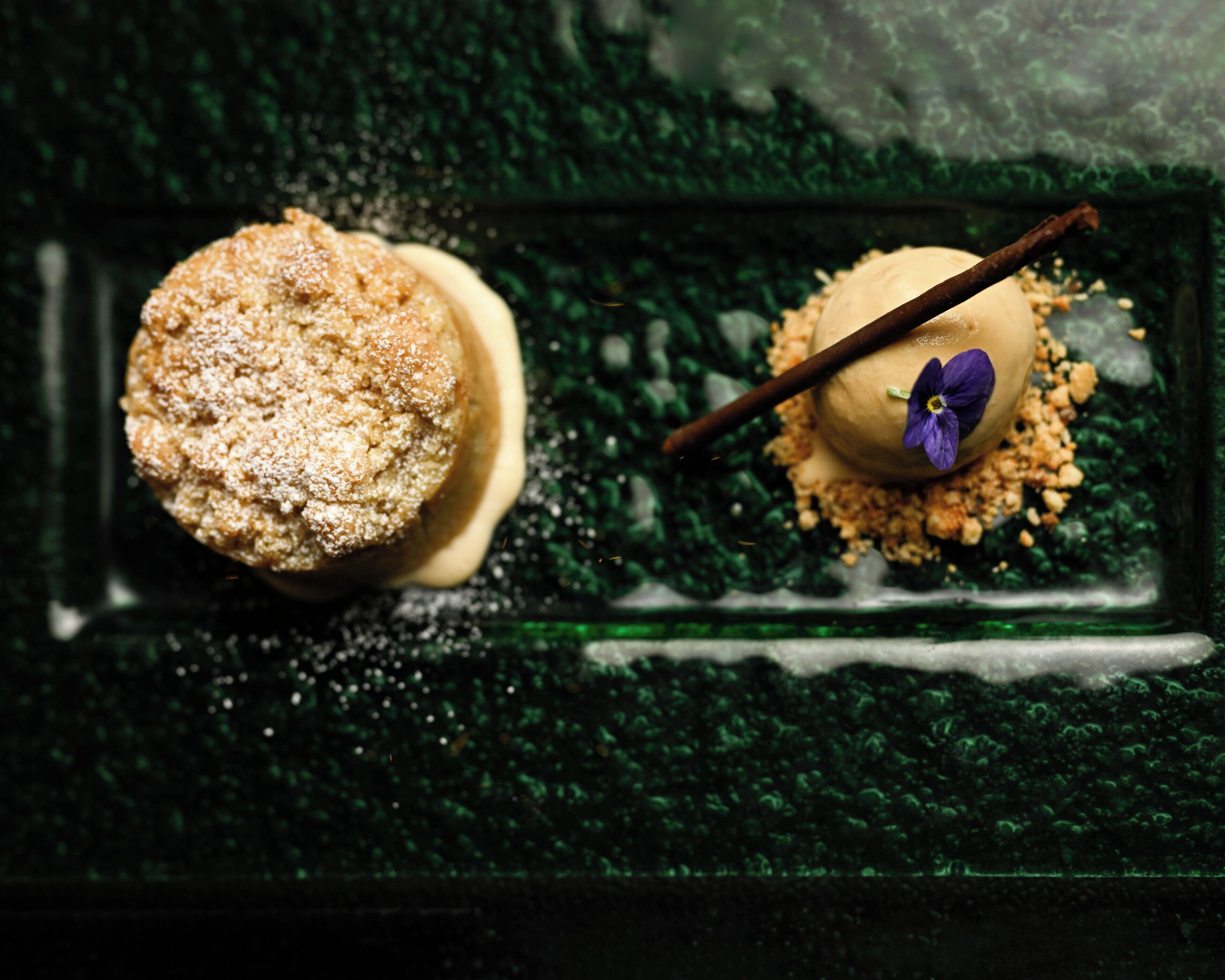 A dessert with a crumbly topping and a flower garnish on a dark, textured surface.