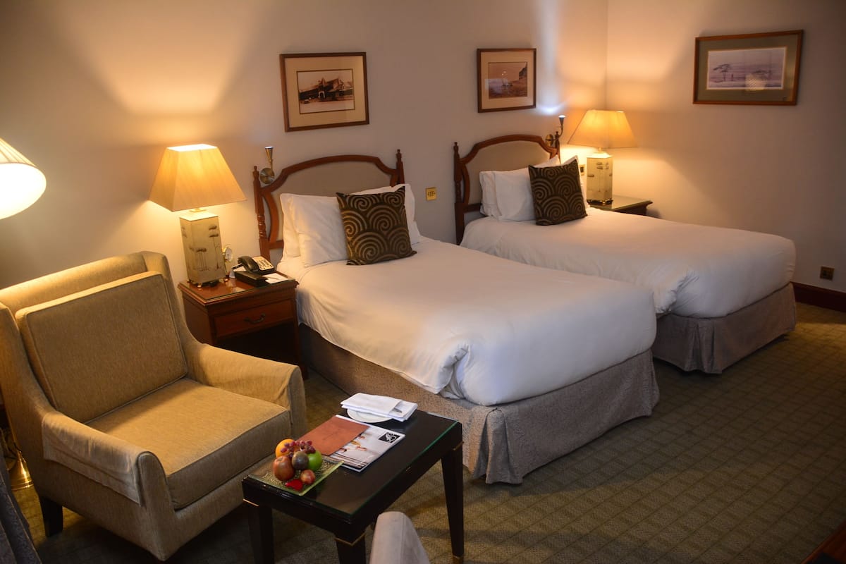 Quarto de hotel com duas camas de solteiro, uma cadeira, uma mesinha de centro com uma fruteira e duas luminárias de cabeceira.