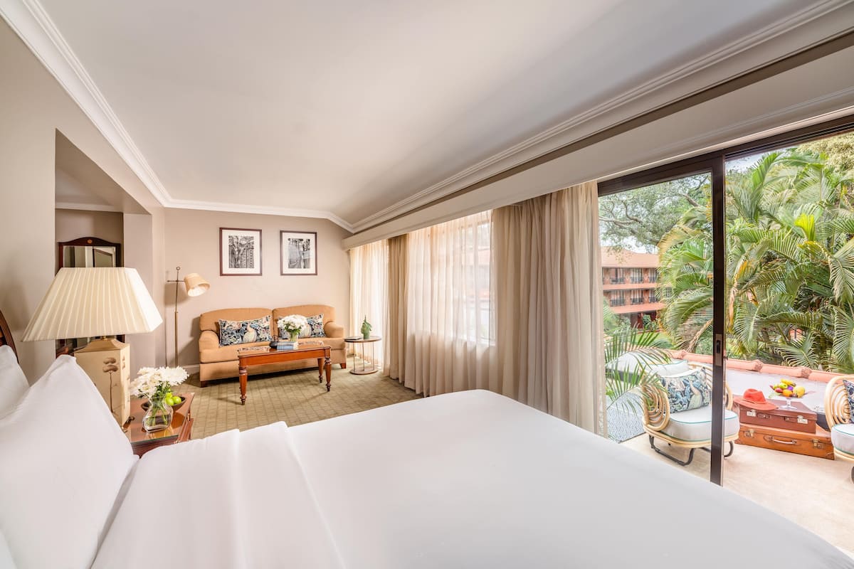 Quarto de hotel com uma cama grande, um sofá, uma pequena mesa com luminária e vista para um jardim tropical através de uma grande janela.