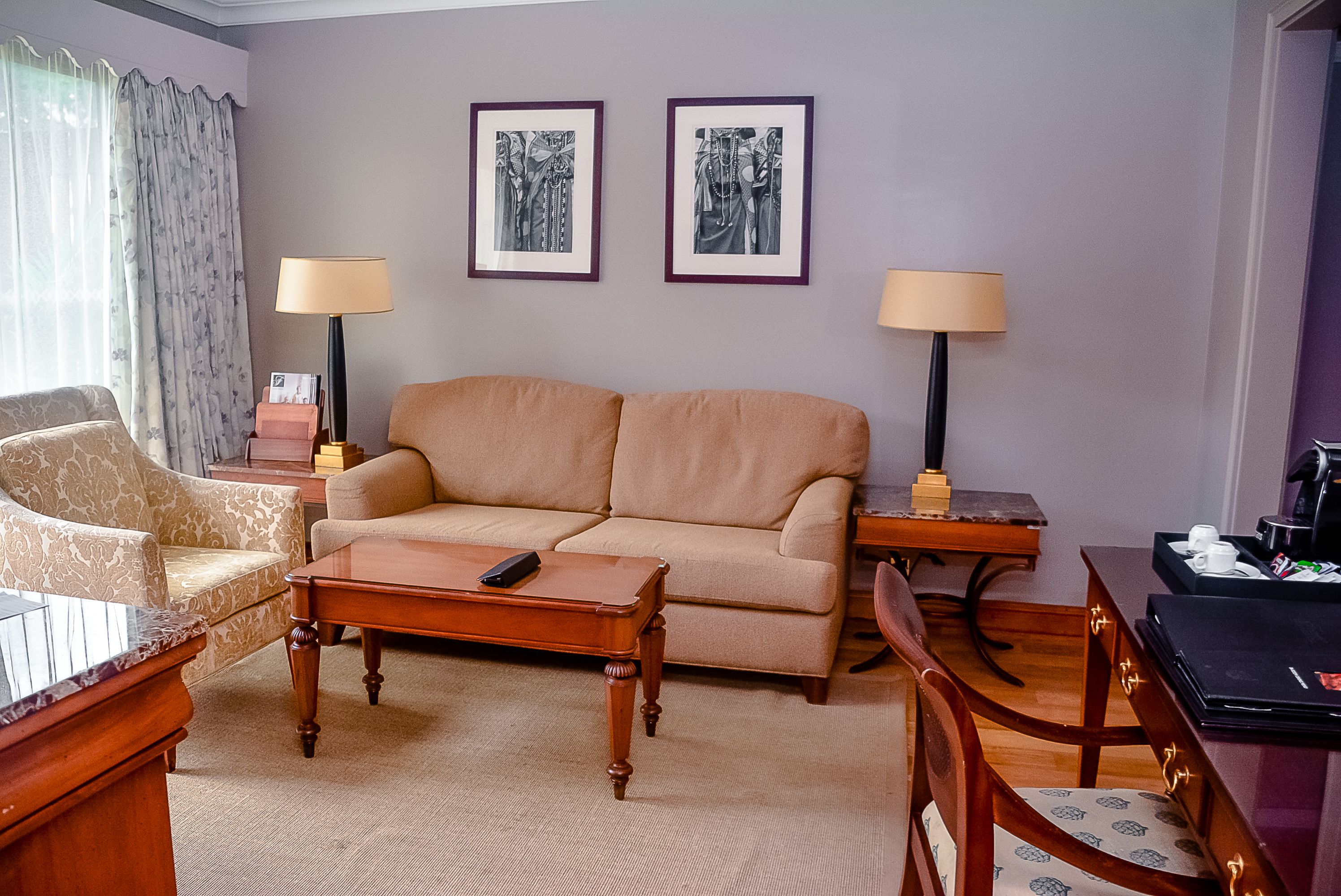 A living room with a beige sofa, two armchairs, a coffee table, a side table with a lamp, and framed artwork on the wall.