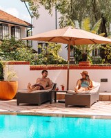 Dos sillones de descanso junto a una piscina con una sombrilla grande, rodeados de vegetación y un área de piscina con azulejos.