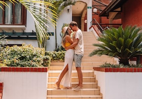 Una pareja abrazándose en una escalera con un muro de ladrillo rojo, plantas de palma y un edificio de fachada roja al fondo.