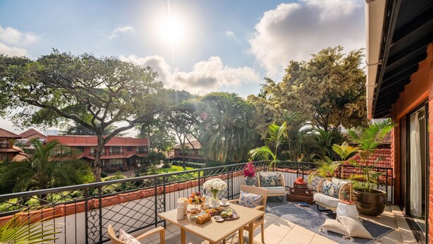A balcony with wicker furniture, a table with fruits and flowers, and a view of surrounding buildings and trees. - Fairmont The Norfolk (Nairobi)