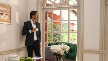 Un hombre con traje sostiene una taza, de pie en una habitación con un sofá verde, una mesa con un jarrón de flores y una computadora portátil.