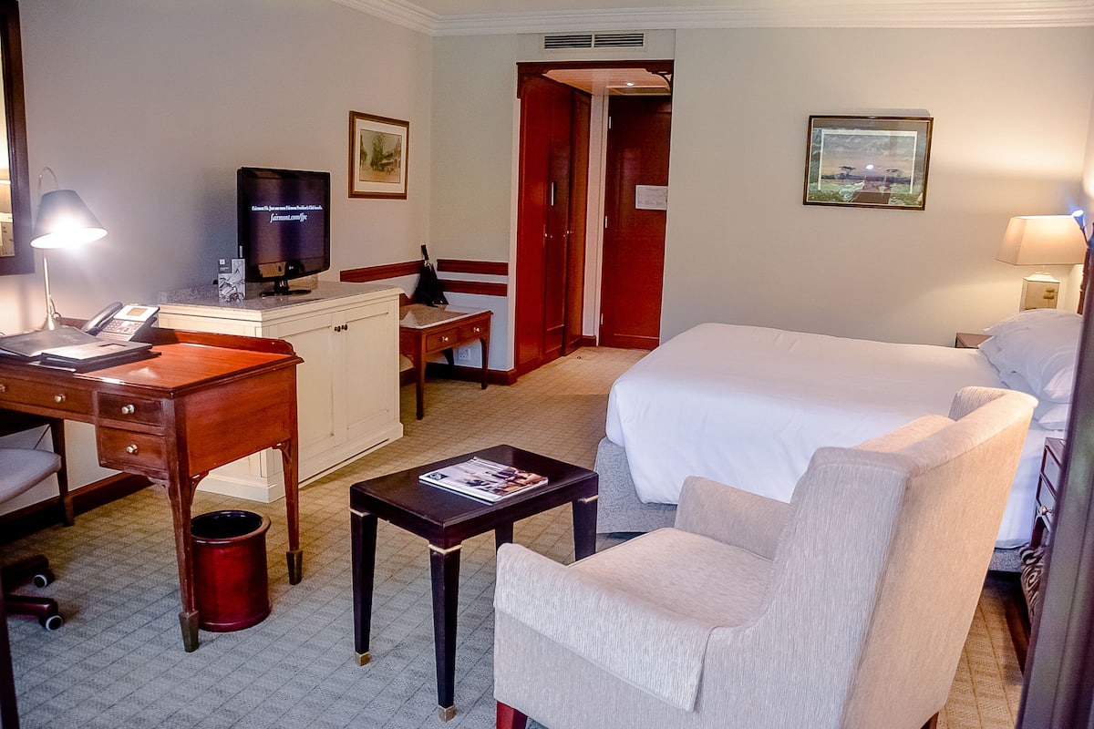 Quarto de hotel com cama, escrivaninha com telefone, televisão, cadeira e pequena mesa com uma revista.