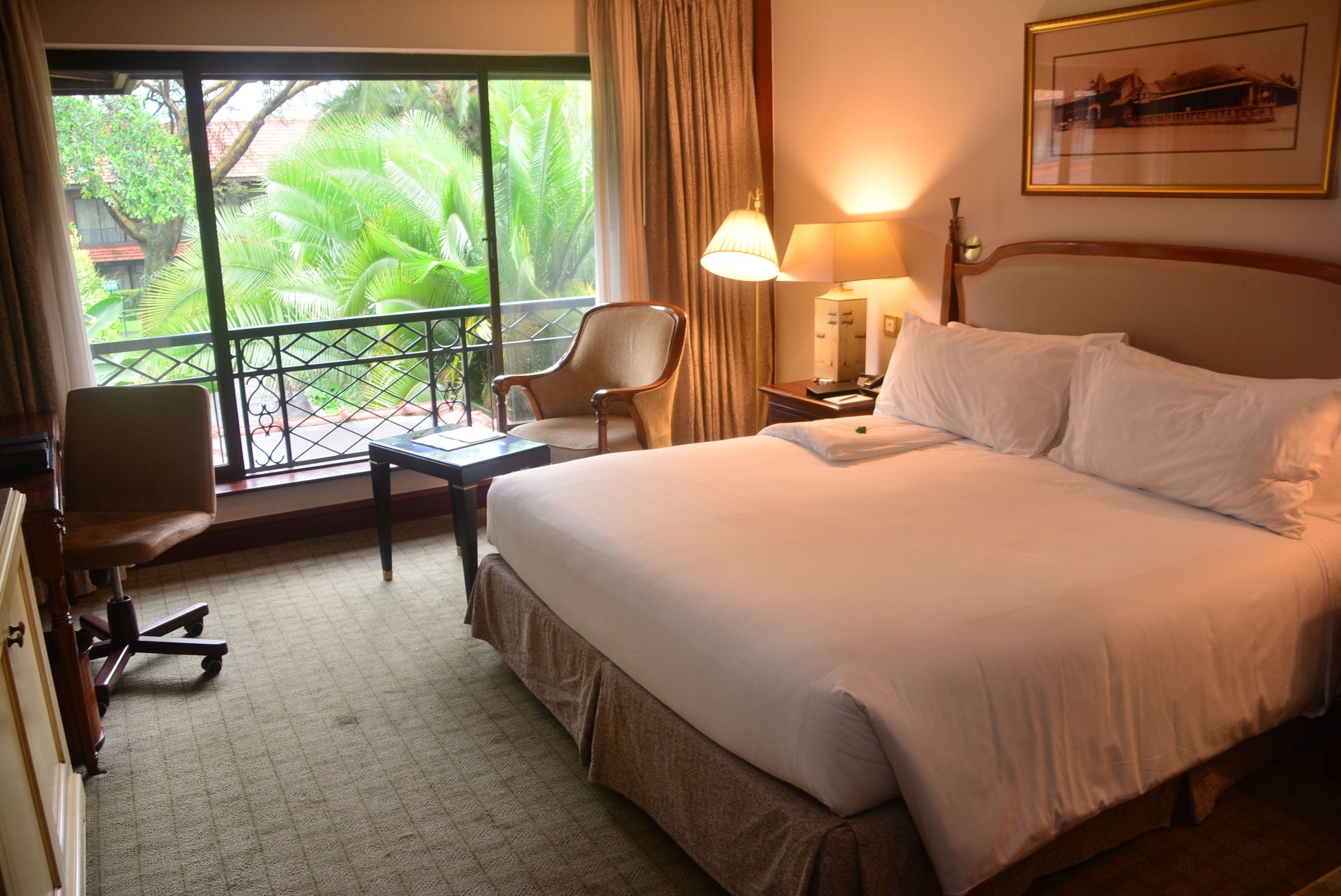 Habitación de hotel con cama, escritorio, silla, lámpara y vistas a zonas verdes por la ventana.