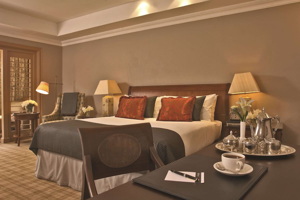 Quarto de hotel com uma cama grande, duas luminárias de cabeceira, um tabuleiro com chá e açúcar, um laptop e uma chávena de café numa mesa.