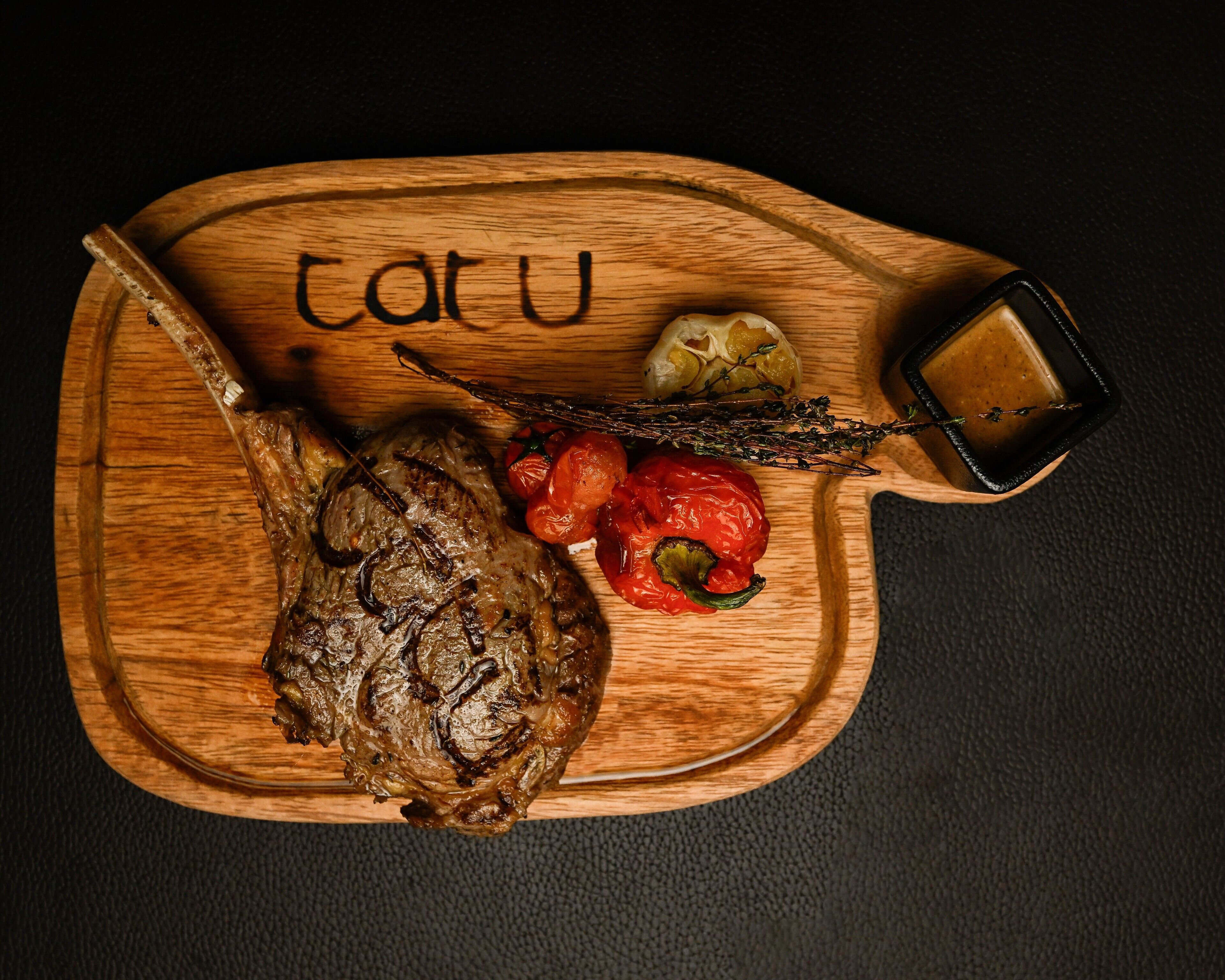 A wooden serving board with a roasted lamb leg, cherry tomatoes, and a sprig of thyme.