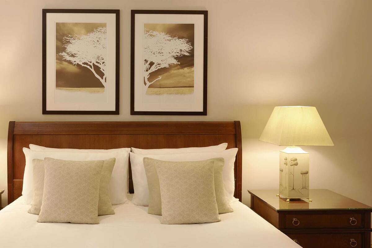 Cama bem arrumada com duas almofadas, flanqueada por duas mesas de cabeceira com abajures, e uma obra de arte na parede acima.