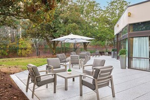An outdoor patio with seating, umbrellas, and a view of trees and a building. - Marriott Raleigh Durham Research Triangle Park (Durham)
