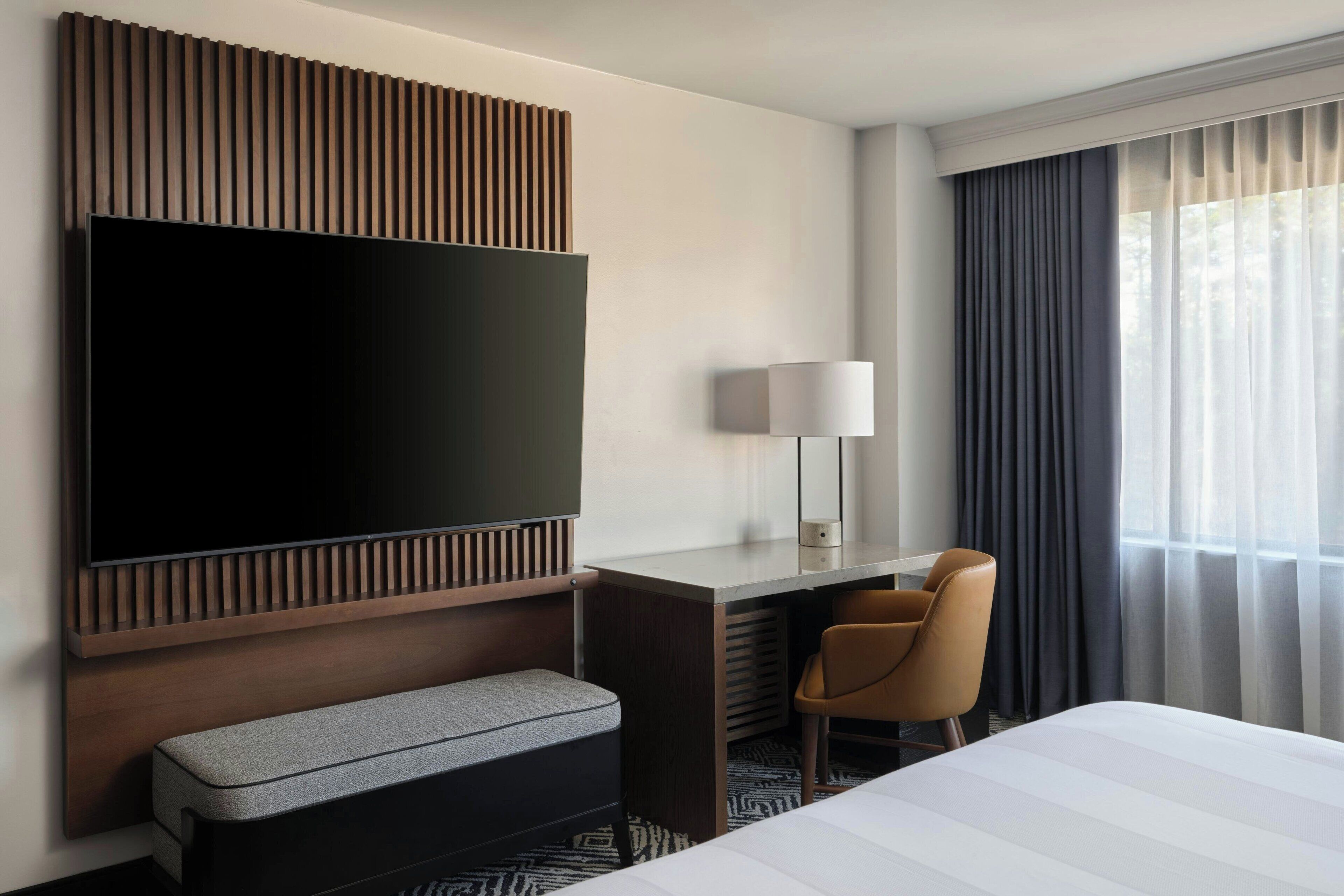 A hotel room with a large flat-screen TV mounted on the wall, a desk with a chair, a bench, and a bed.