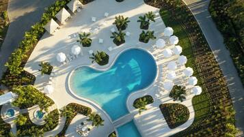 An aerial view of a resort with a heart-shaped pool, surrounded by lounge areas with umbrellas and chairs, and palm trees.