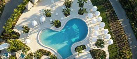 Ovenfra ses et resort med en pool i hjerteform, omgivet af loungeområder med parasoller og stole, samt palmer.