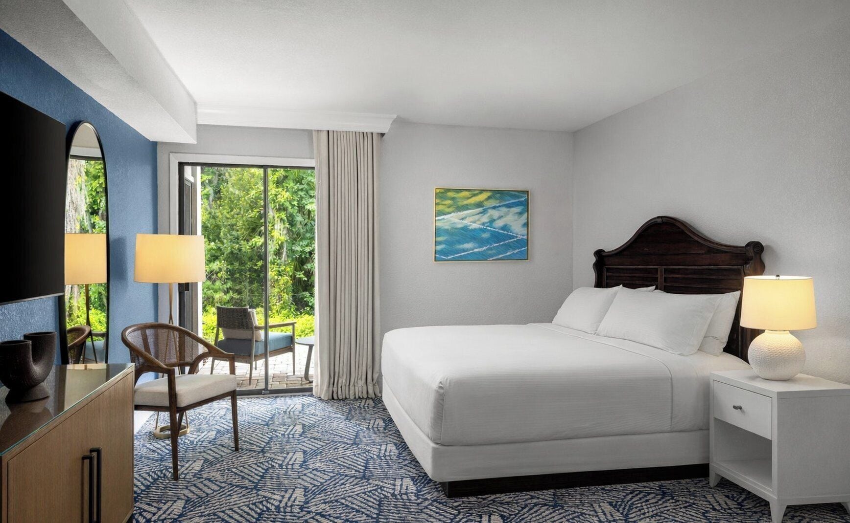 A hotel room with a bed, bedside table, lamp, chair, and a view of greenery through the window.