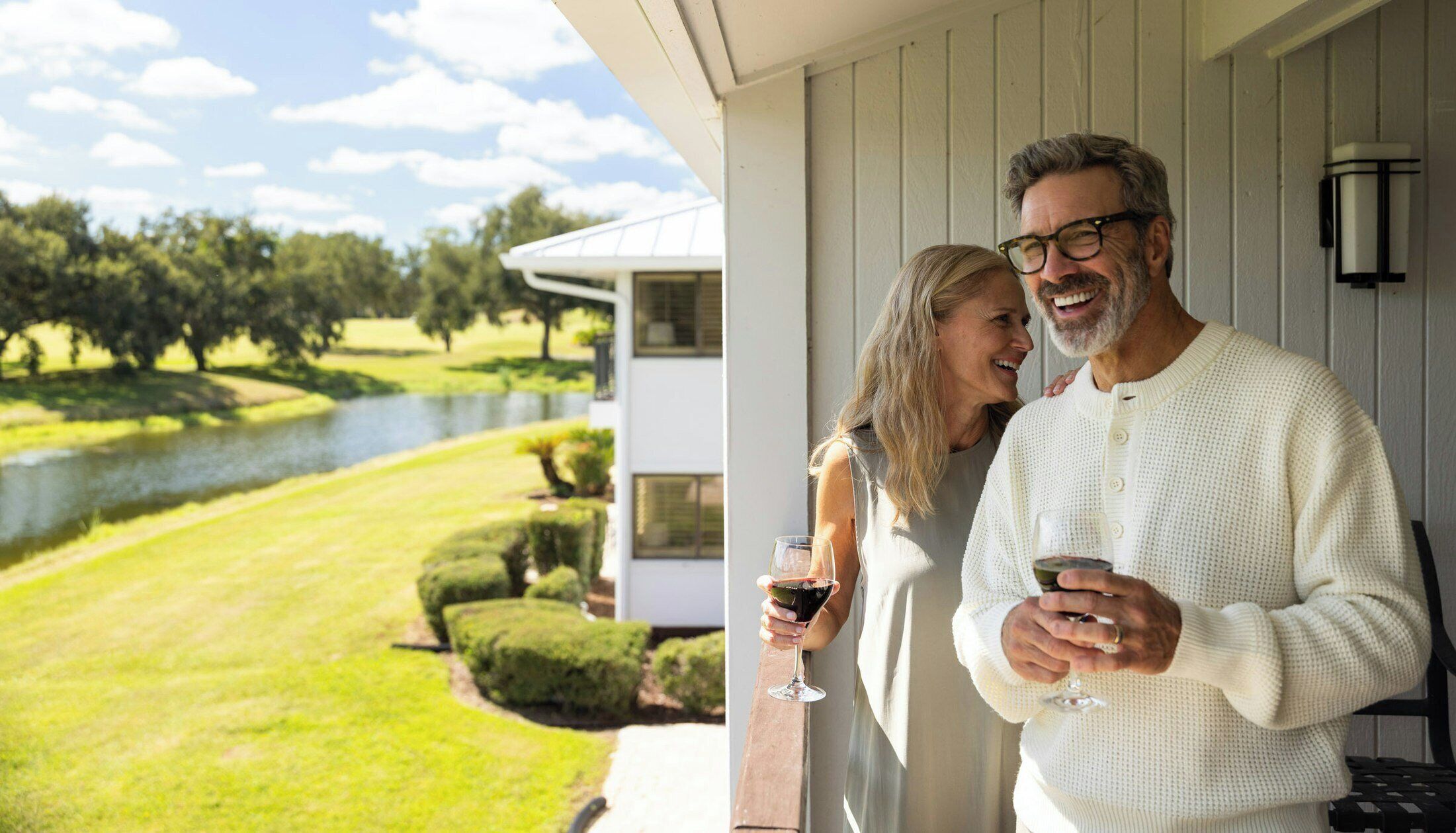 A couple standing on a porch with a view of a lake and manicured lawn.
