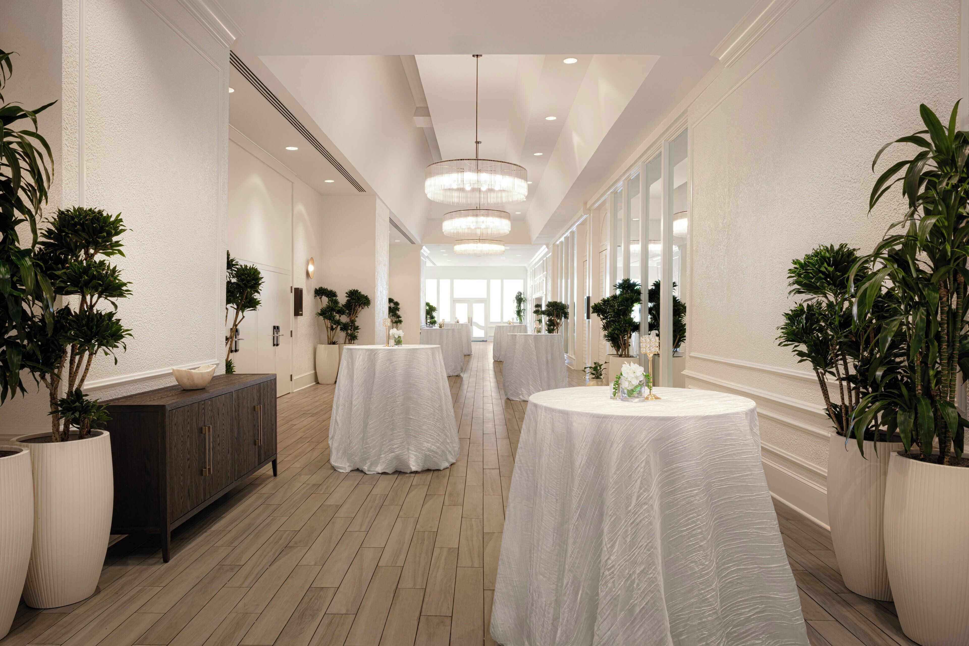 A hotel corridor with round tables covered in white tablecloths, flanked by potted plants and a wooden cabinet.