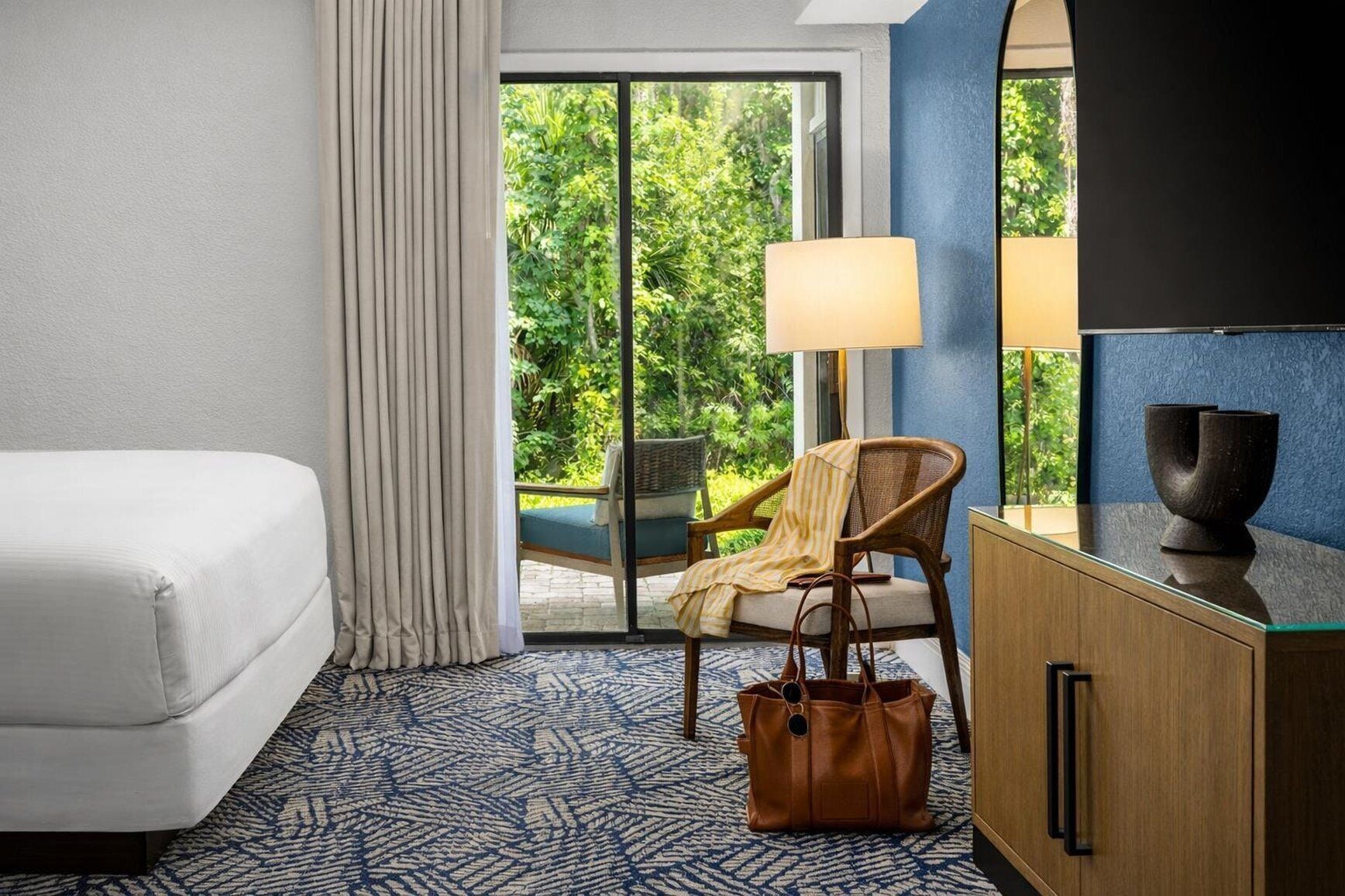 Quarto de hotel com cama, cadeira, escrivaninha, abajur e vista para o verde pela janela.