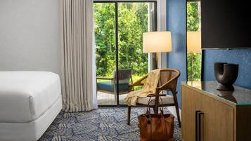 Quarto de hotel com cama, cadeira, escrivaninha, abajur e vista para o verde pela janela.
