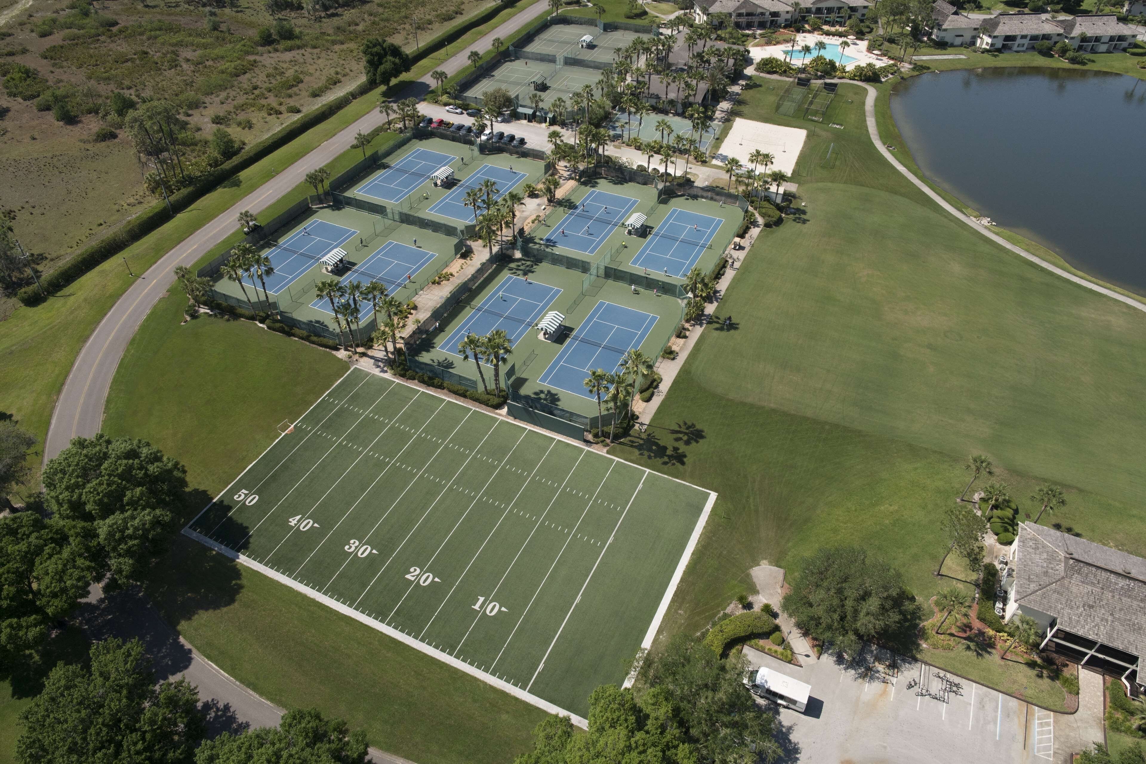 Une vue aérienne d’un complexe sportif comprenant plusieurs courts de tennis, un terrain de football et un lac.