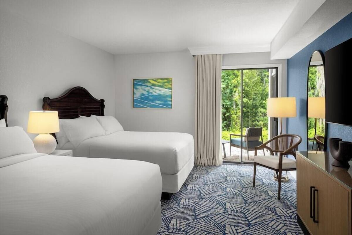 Ein Hotelzimmer mit zwei Betten, einem Schreibtisch, einem Sessel und einem großen Spiegel, mit Blick auf Grünflächen.