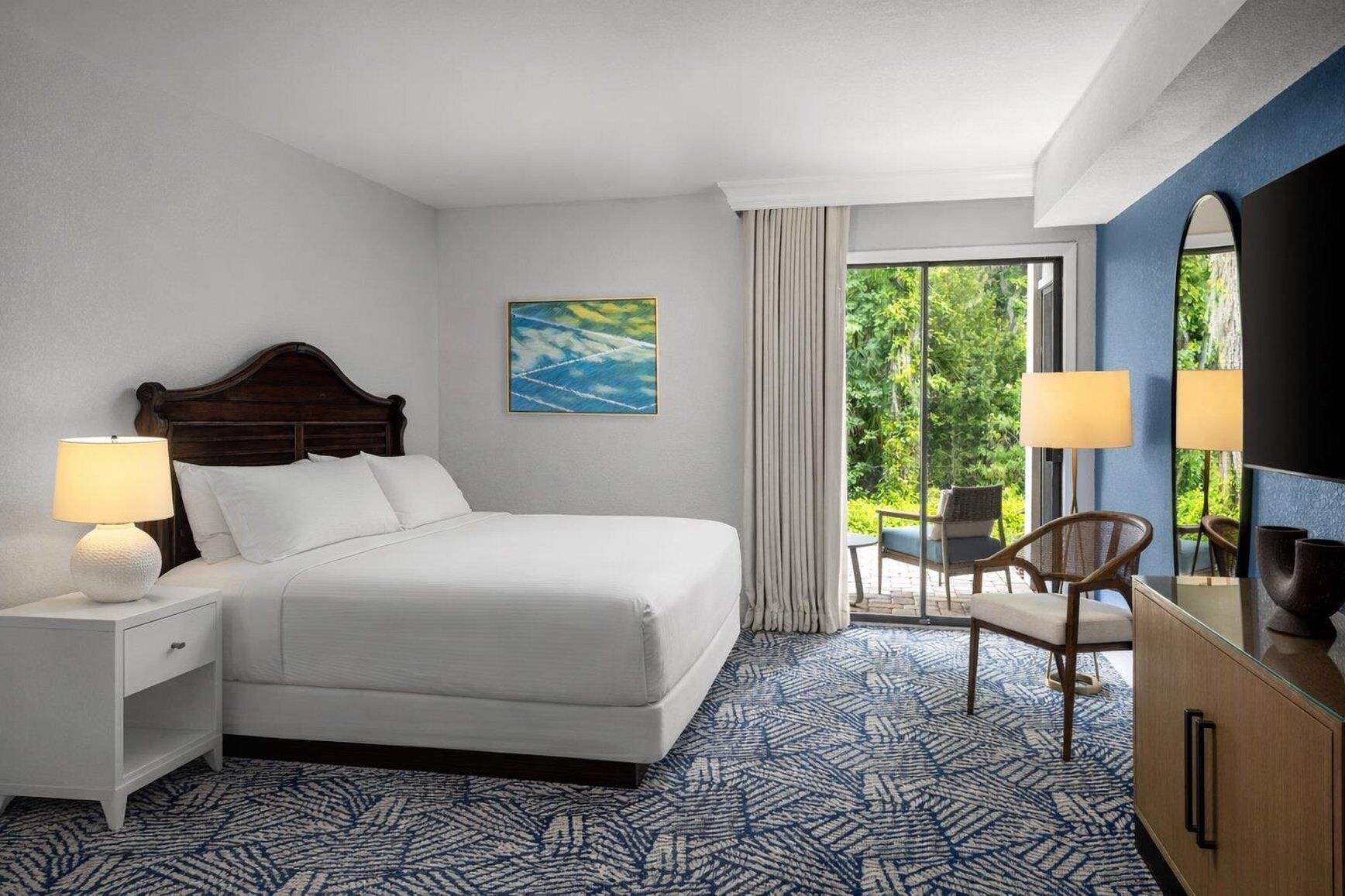 Une chambre d’hôtel avec un lit, deux tables de chevet, une chaise, un téléviseur et une vue sur la verdure par la fenêtre.