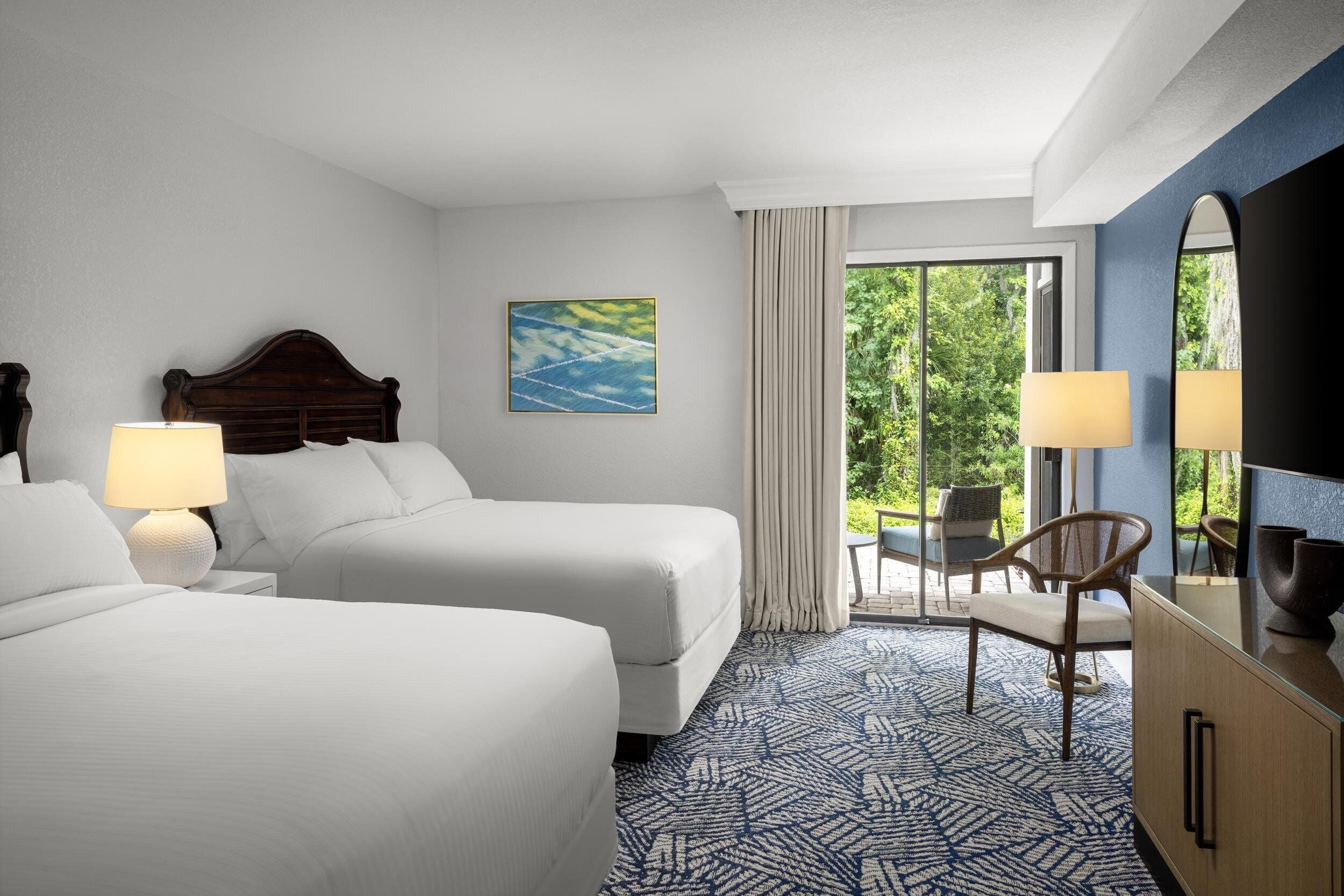A hotel room with two beds, a desk, a chair, a large mirror, and a view of greenery outside.