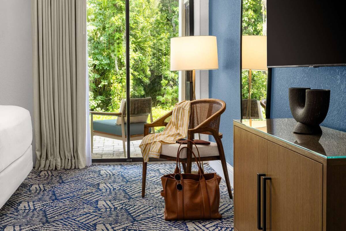 Ein Hotelzimmer mit Bett, Stuhl, Schreibtisch, Lampe und Blick auf Grünflächen.