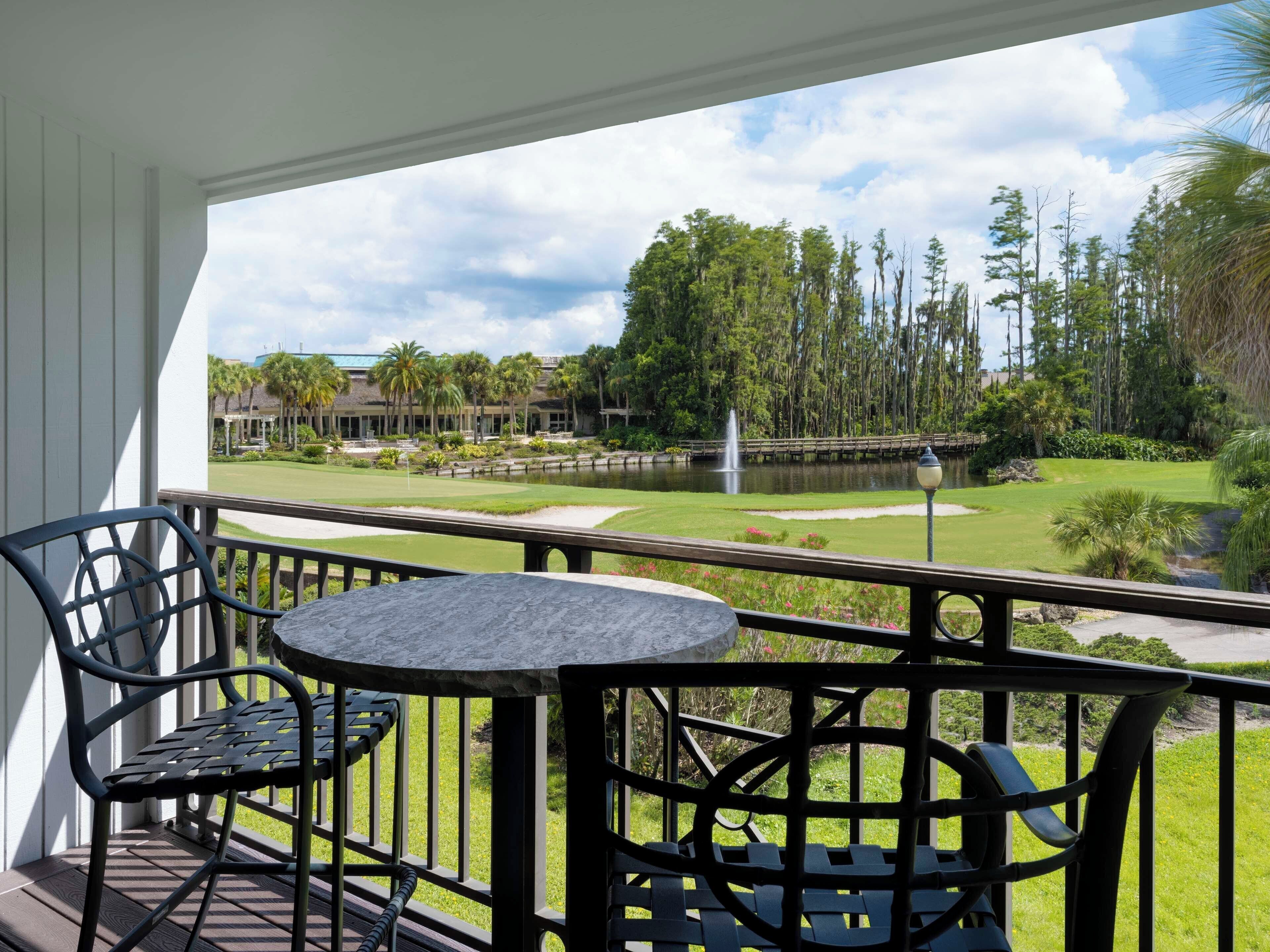 A balcony with a table and chairs, overlooking a golf course with a pond and trees.