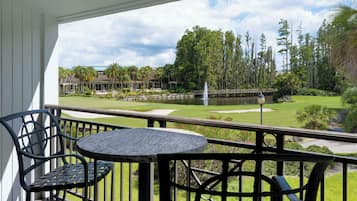 A balcony with a table and chairs, overlooking a golf course with a pond and trees.