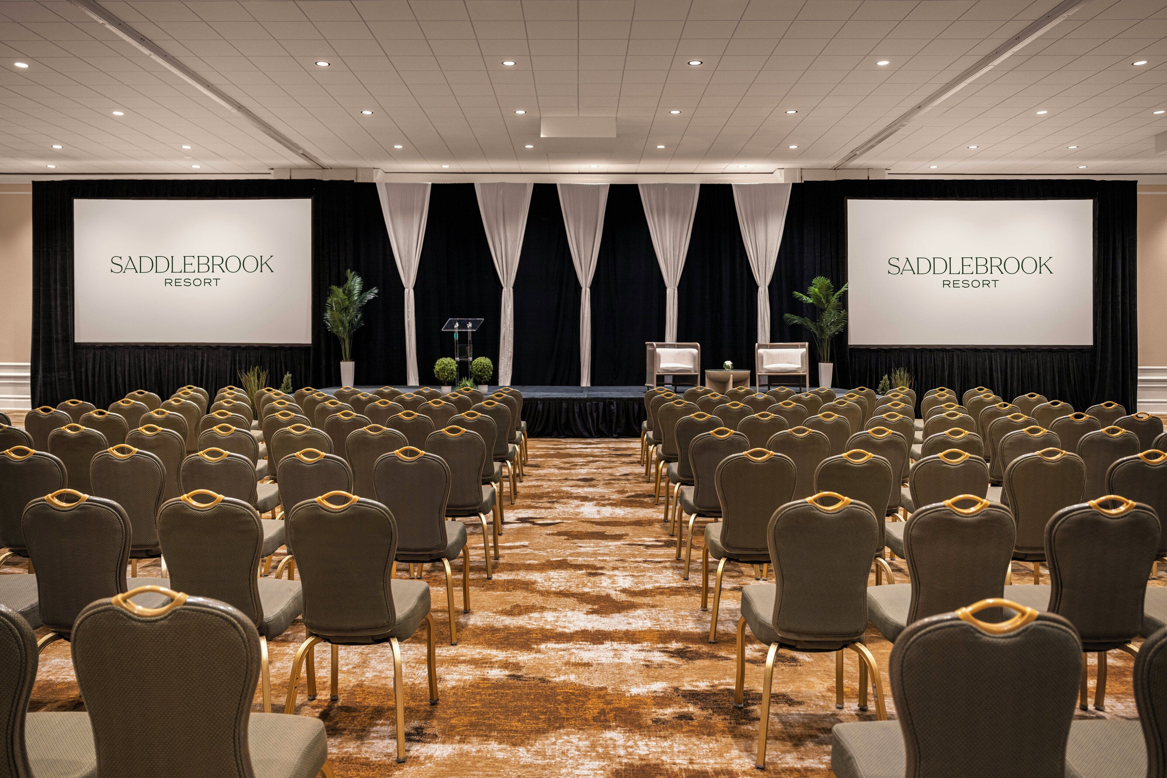 Un salón de conferencias con filas de sillas, un escenario con una pantalla mostrando 'SADDLEBROOK RESORT', y suelo alfombrado.