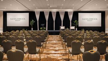 Un salón de conferencias con filas de sillas, un escenario con una pantalla mostrando 'SADDLEBROOK RESORT', y suelo alfombrado.