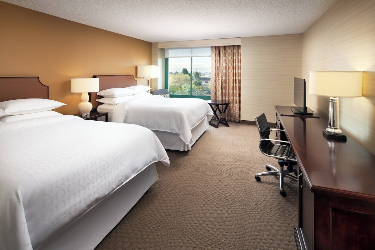 A hotel room with two beds, a desk, a chair, a TV, and a window with curtains.
