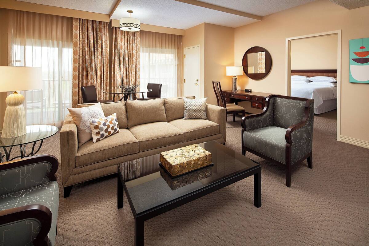 A hotel room with a beige sofa, a matching armchair, a coffee table, a desk with a lamp, a bed, and a large window with curtains.