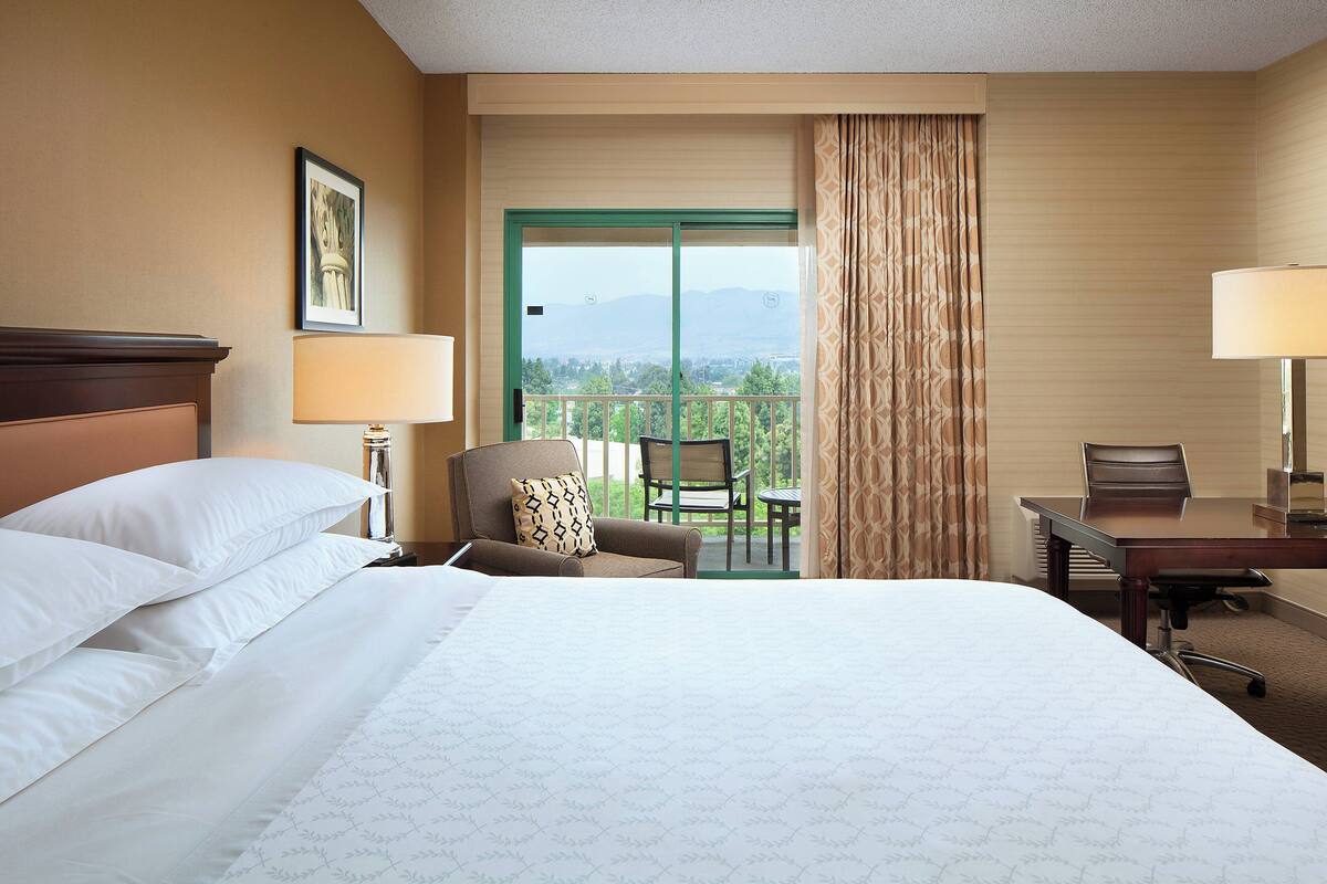 A hotel room with a large bed, a desk, a chair, a balcony with a view of the mountains, and a window with curtains.