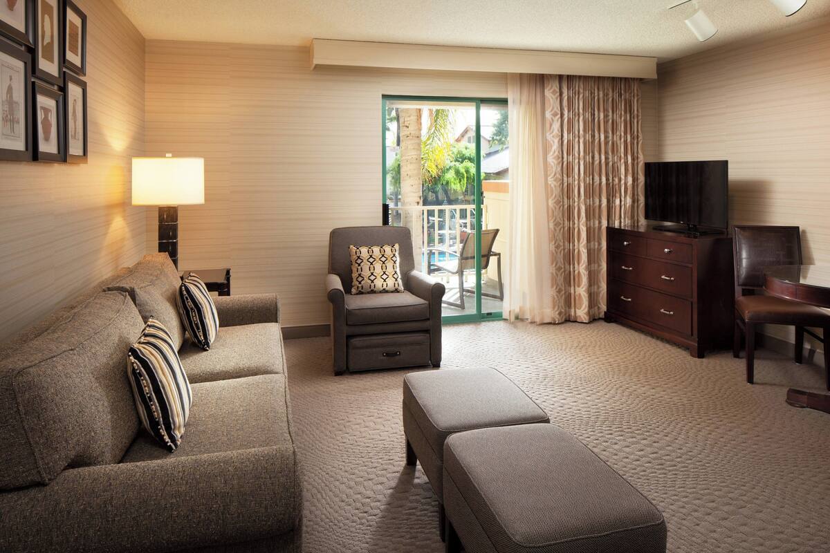 A hotel room with a sofa, ottoman, armchair, TV, and a sliding glass door leading to a balcony.