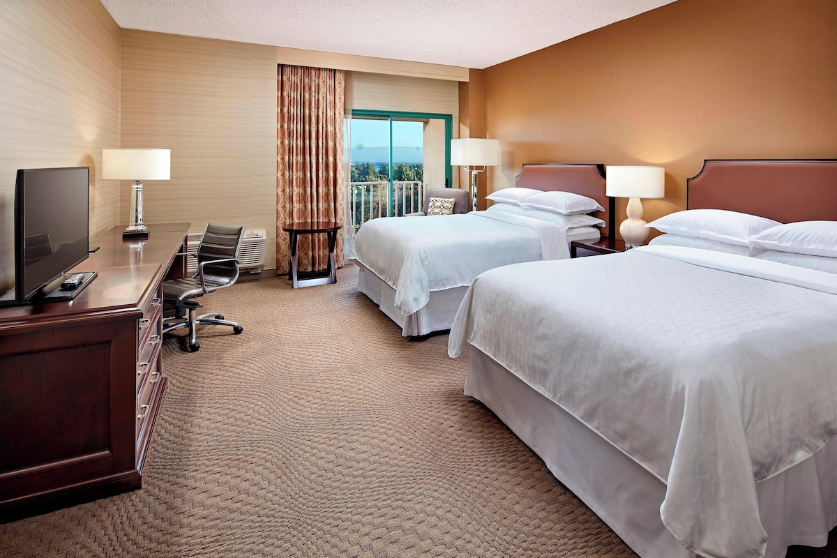 A hotel room with two beds, a desk with a TV, and a view of the outdoors.