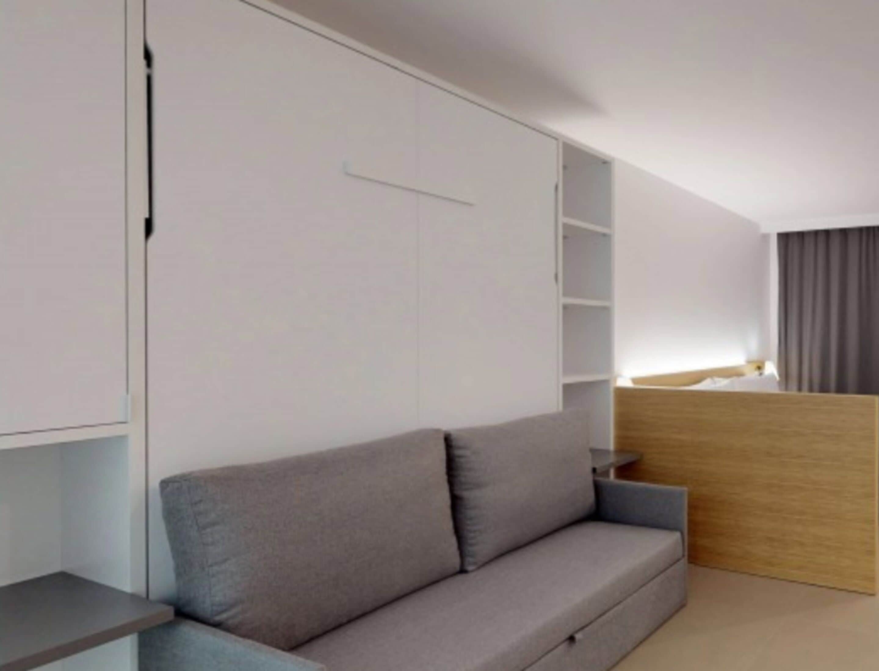 A modern hotel room with a grey sofa, a wooden desk, and built-in shelving.