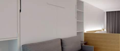 A modern hotel room with a grey sofa, a wooden desk, and built-in shelving.