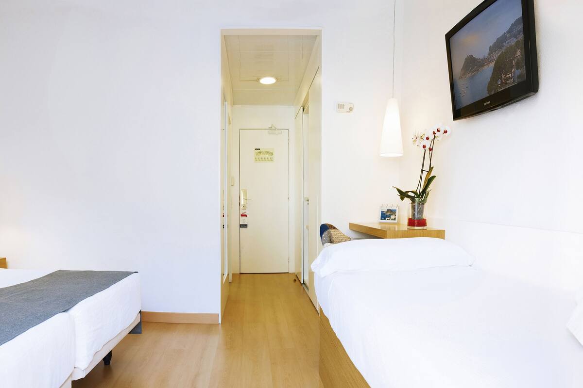 Een hotelkamer met twee bedden, een lijst met een afbeelding van een zeilboot, een bedlamp, een kleine tafel met een bloevas, en een televisie die aan de muur is bevestigd.