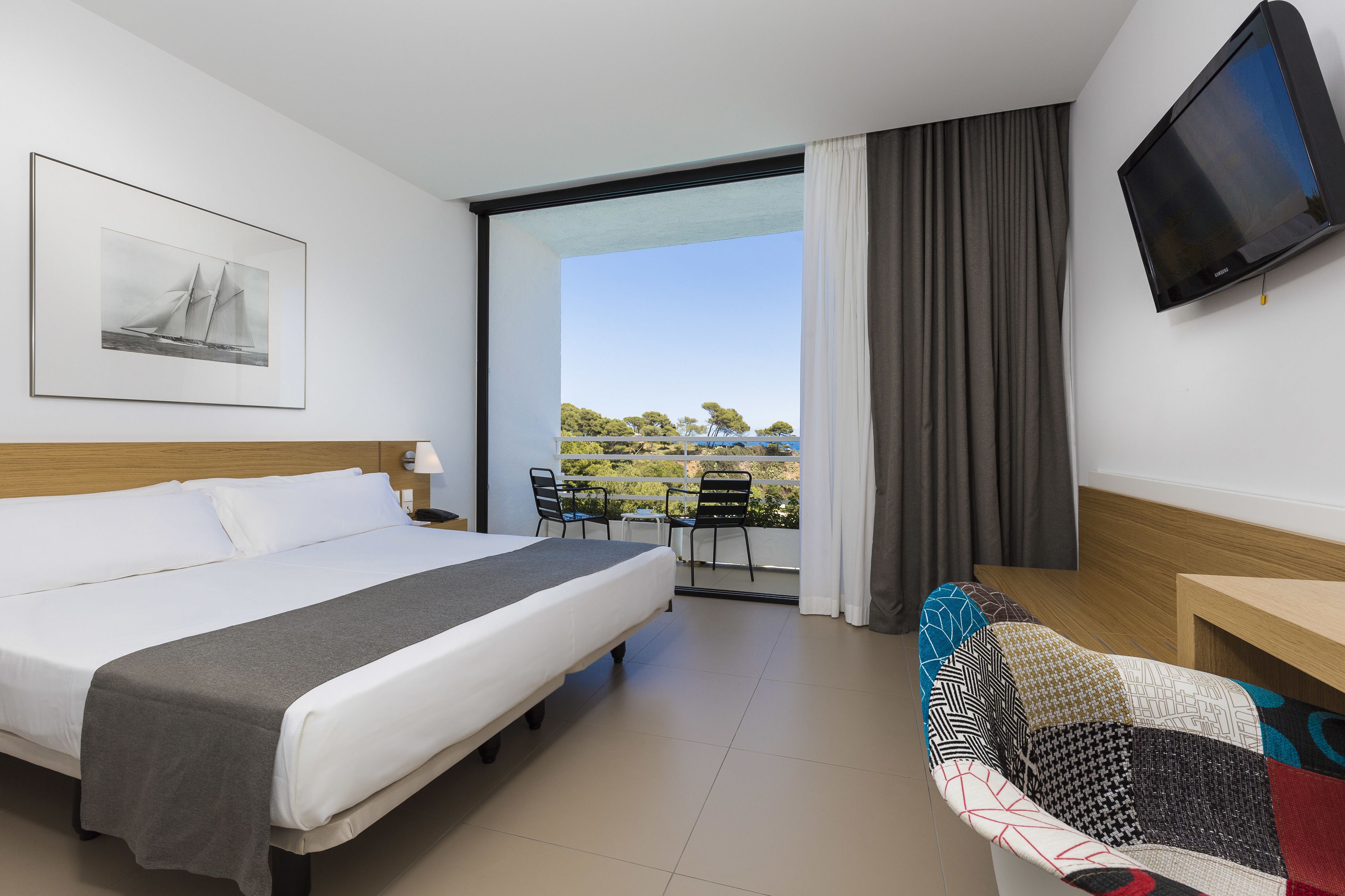 Habitación de hotel con cama, televisión, balcón con sillas y un cuadro en la pared.