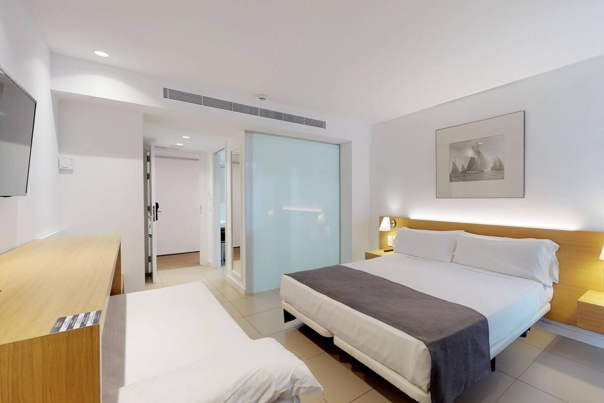 A hotel room with a bed, bedside tables, a desk, a wardrobe, and a door leading to another room.